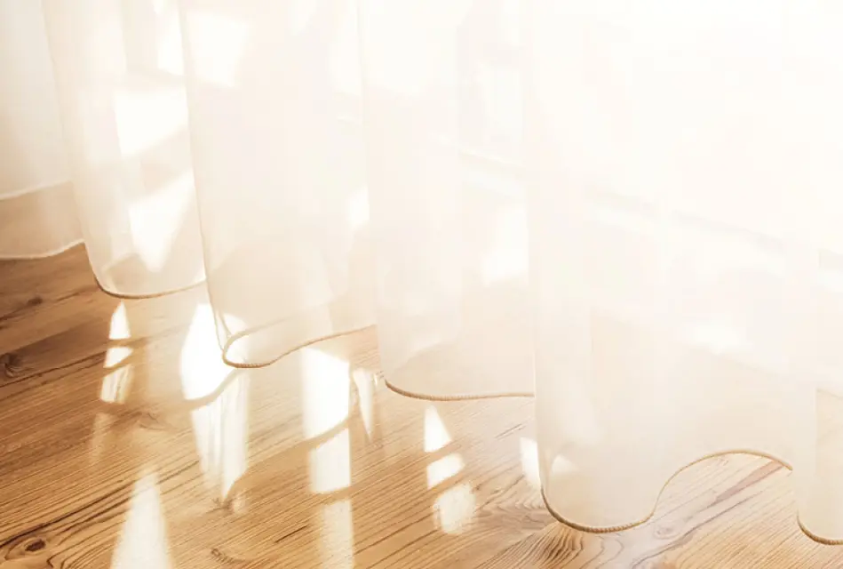 Transparente weiße Gardinen vor Fenster, Sonnenlicht fällt auf hellen Holzboden und erzeugt Schattenmuster